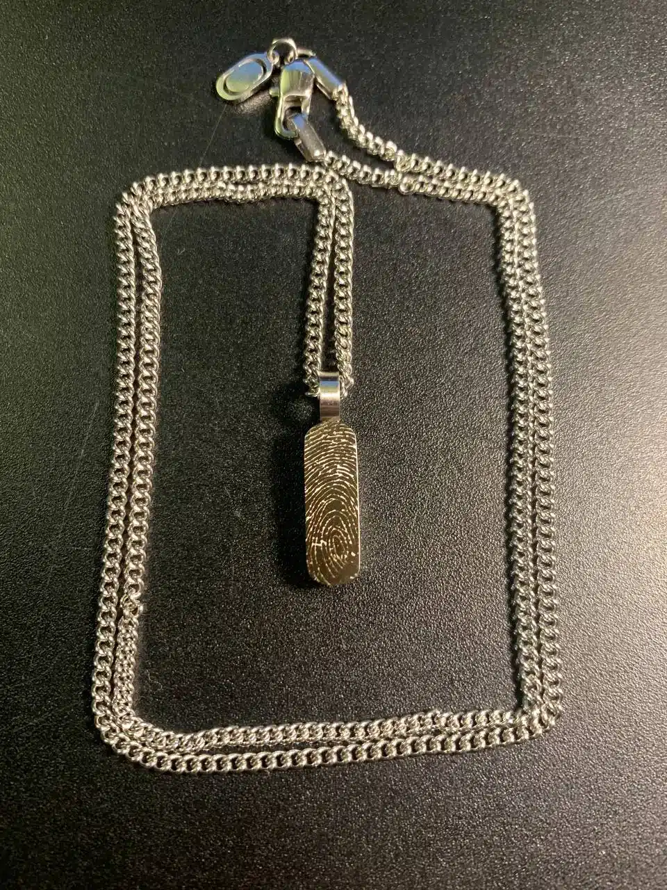 Sormenjälkikaiverrus metalliriipukseen 3 Metalliriipus sormenjälkikaiverruksella ja ketjulla tummalla pöytäpinnalla.